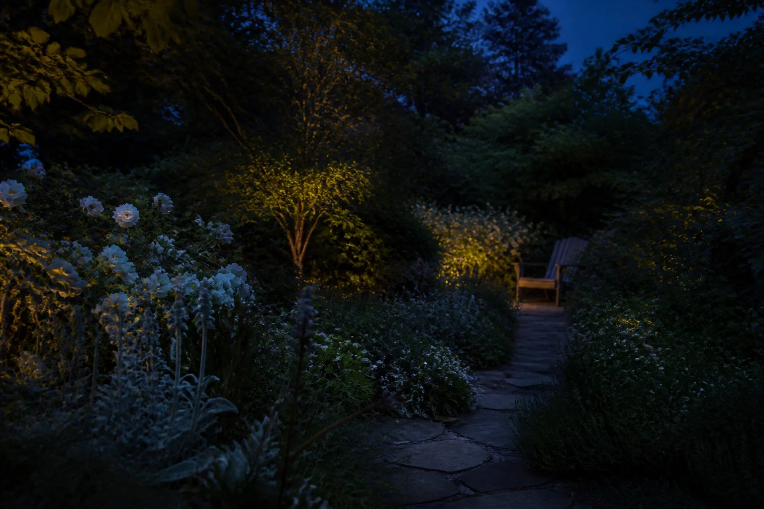 A stone path passes pale flowers and silvery foliage toward a wooden chair in a deep blue garden after sunset.