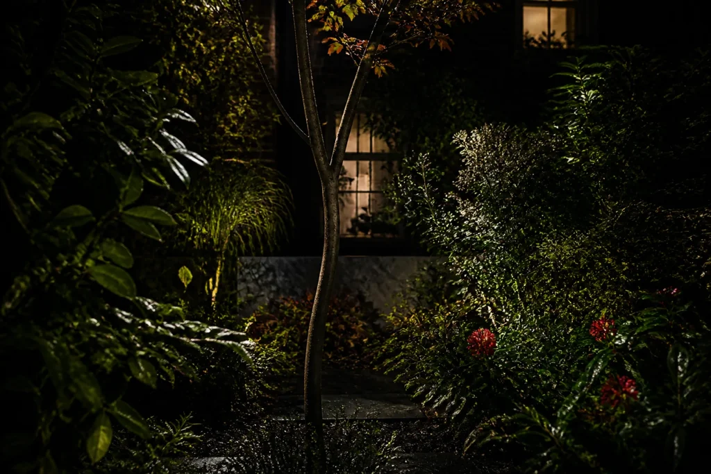 Layered shrubs and a slim tree interrupt views of lit neighboring windows in a dark urban garden.