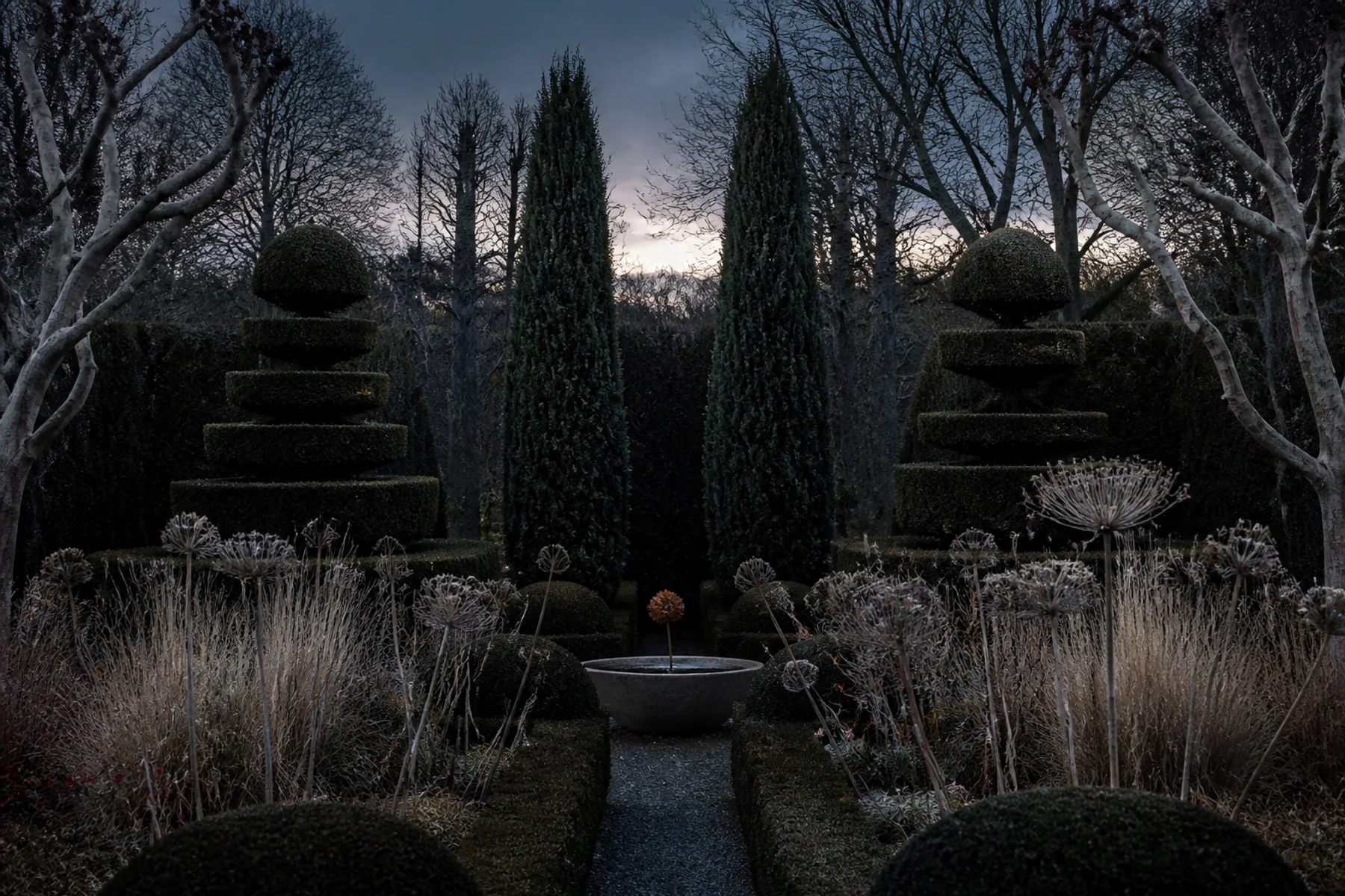 A winter garden at twilight with clipped dark evergreens, bare branching trees, pale seedheads, and a narrow gravel path.